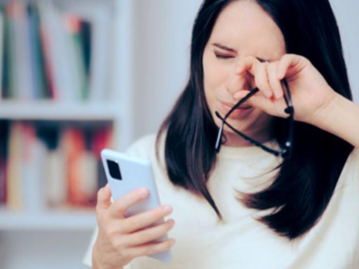 A picture that shows a woman in her 30s looking at her phone while rubbing her eyes.