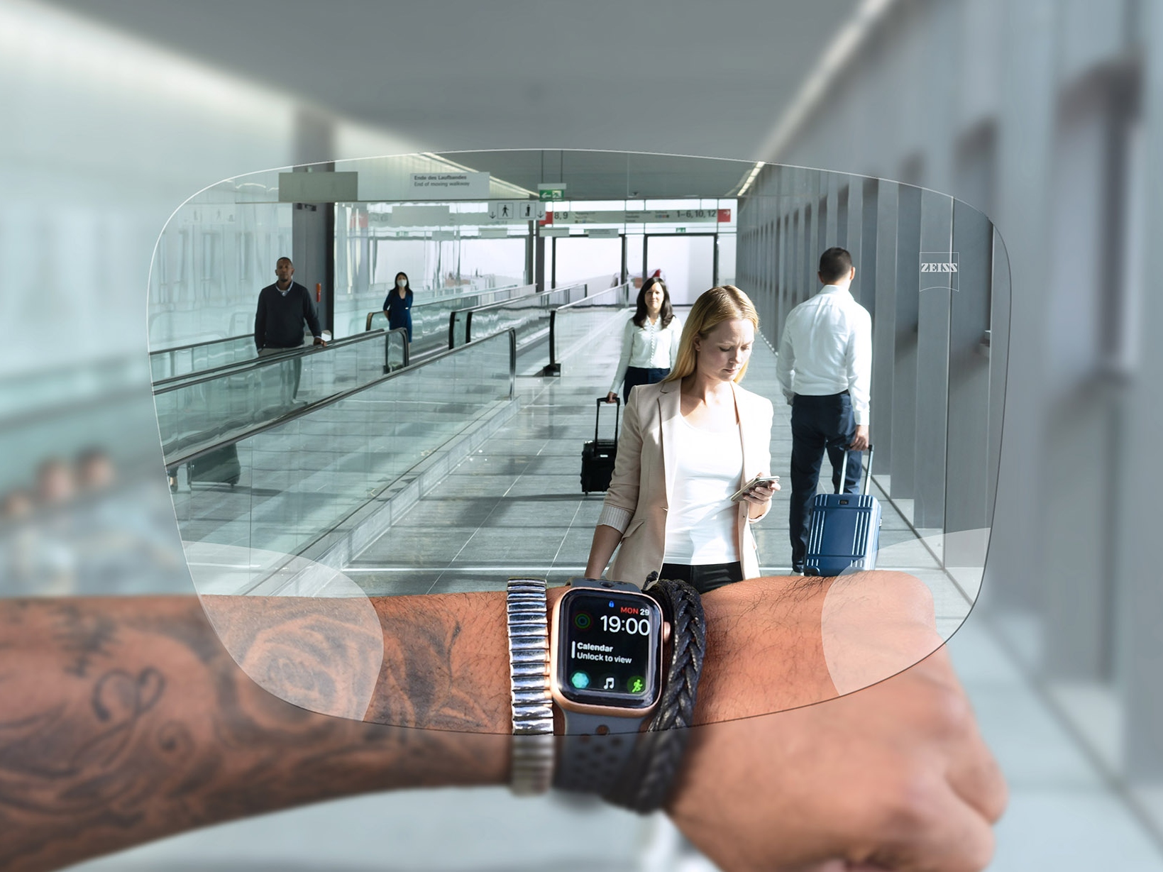 Person looks at his watch through ZEISS SmartLife Digital lenses. In the background you see a typical airport situation with people walking on escalators and pushing their suitcases. 